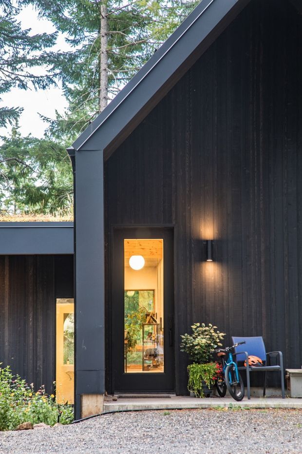 Exterior view of a dark, steeply pitched gable-roofed building surrounded by mature trees. The facade is clad in near-black vertical timber, with charcoal-grey metal fascia and trim. A narrow glazed entry door reveals a warmly lit interior, while wall-mounted lighting, potted plants, a bicycle and outdoor seating sit near the entrance. Gravel paving and low garden planting complete the approach.