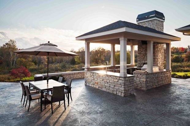 A spacious outdoor entertaining terrace featuring a stone-clad pavilion with a hipped roof, chimney, built-in fireplace, and open sides supported by square columns. A dining table with chairs and a large umbrella occupies the paved terrace, overlooking landscaped surroundings.
