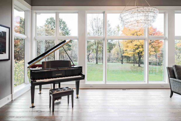 Bright, elegant music room or living area featuring a black grand piano and matching bench positioned against expansive white-framed windows. The room has pale timber flooring, light walls, a dark ceiling, a contemporary crystal chandelier, and a view onto a leafy landscaped property with autumn trees. A partial lounge chair and framed artwork are visible at the edges.