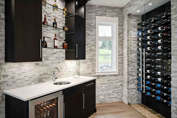 A compact, highly finished wine room with dark built-in cabinetry, open bottle display racks, a white stone-topped wet bar with sink, under-counter beverage refrigerator, and a full-height light grey stacked-stone feature wall. A window provides natural light, while recessed ceiling lighting illuminates the room.