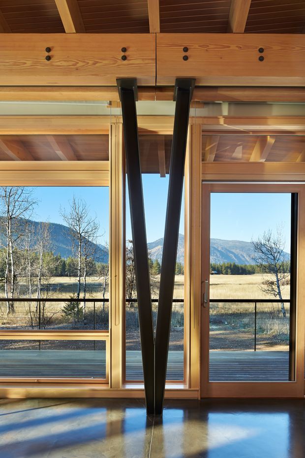 Interior architectural view dominated by exposed timber roof framing, warm wood beams and trim, expansive timber-framed glazing, and a prominent black V-shaped structural support. The glazing opens onto a deck and a clear mountain valley landscape. A polished concrete floor reflects daylight and the structural elements.
