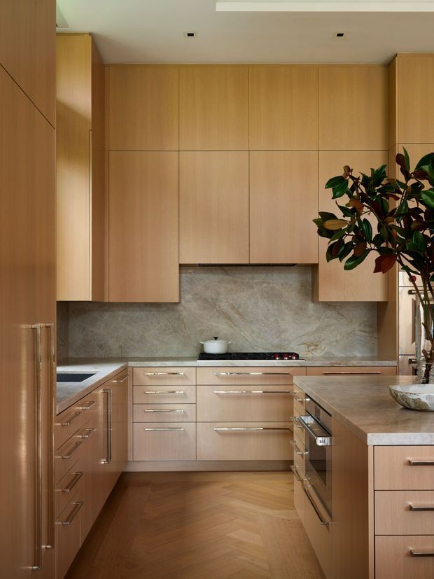 A warm, refined contemporary kitchen with extensive light timber cabinetry, integrated upper storage, a pale natural-stone backsplash, and a timber-look island with drawers and built-in appliances. The floor is laid in a herringbone pattern, while a large leafy indoor plant adds organic contrast. Recessed ceiling lighting and clean flush cabinetry create a streamlined, highly functional composition.