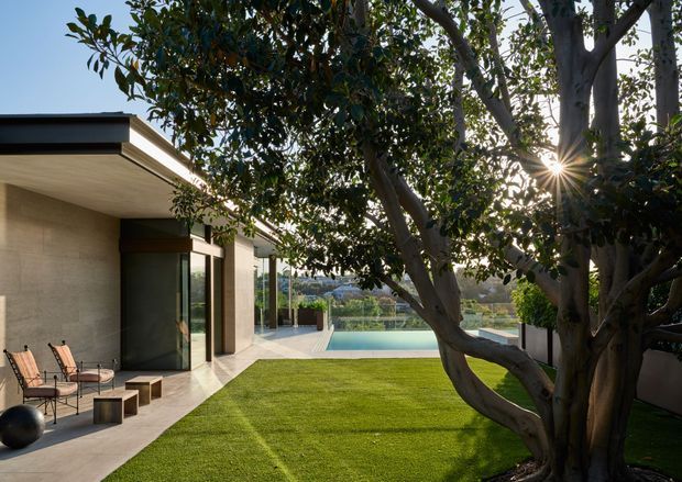 A contemporary single-storey residence viewed from a landscaped backyard, with a broad low-pitched roof, textured neutral walls, expansive glazed doors, a paved terrace with outdoor seating, manicured lawn, mature tree, and a rectangular swimming pool beyond.