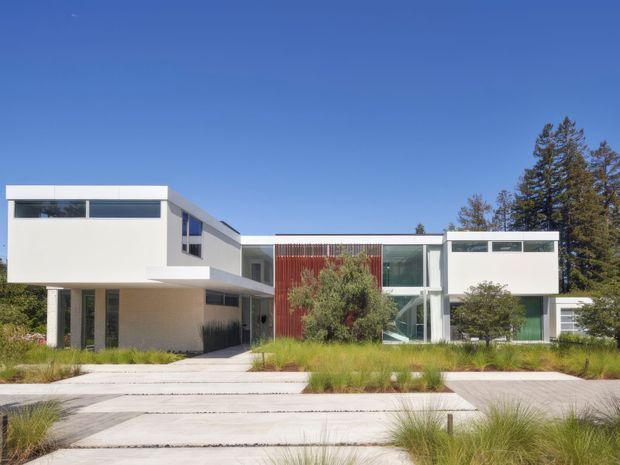 Exterior view of a large contemporary two-storey residence with crisp white rendered volumes, expansive horizontal glazing, a prominent glazed central section, and a vertical reddish timber screen. The house is set behind a landscaped front garden with ornamental grasses and mature trees, approached by broad pale concrete paving under a clear blue sky.