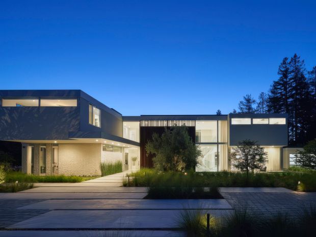 Twilight exterior view of a large contemporary multi-storey residence with crisp rectilinear volumes, dark and light facade sections, extensive illuminated glazing, a sheltered entry, and landscaped planting. Broad paved areas and a linear water feature or reflective surface occupy the foreground, with mature trees behind the home.