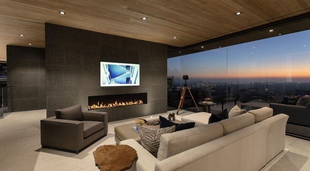 Luxurious contemporary living room with a large cream sectional sofa and dark upholstered armchair arranged around a long linear gas fireplace. The fireplace wall is clad in dark textured stone or tile and incorporates a wall-mounted screen. A warm timber-lined ceiling with recessed downlights extends to full-height corner glazing, overlooking a city skyline at dusk. The room has a neutral, refined palette with layered geometric cushions and a sculptural natural wood side table.