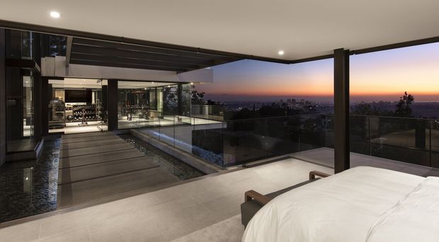 A luxurious modern bedroom opening onto a broad covered terrace or balcony at dusk. The foreground shows a neatly made bed, while frameless glass balustrades, dark structural columns, large glazed openings, and a linear outdoor water feature or pool create a seamless indoor-outdoor connection. A panoramic city and sunset view extends beyond the terrace.