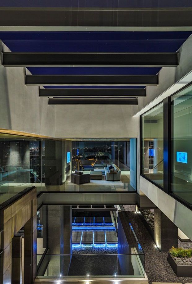 Interior architectural view of a dramatic modern multi-level space organized around a glazed internal void or courtyard. Exposed concrete walls, dark steel beams, extensive black-framed glazing, reflective dark surfaces, and blue-toned concealed lighting create a luxurious, moody atmosphere. A living area with sofas is visible beyond the glass, while a lower-level reflective water or skylight feature is visible below.