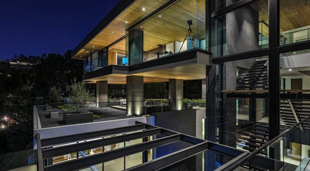 Night-time exterior view of a multi-level contemporary residence with extensive floor-to-ceiling glazing, dark metal framing, warm timber soffits, cantilevered volumes, and illuminated landscaped terraces. An internal stairway and parts of the interior are visible through the glass. The design features strong horizontal and vertical geometry, recessed lighting, and a flat-roof form.