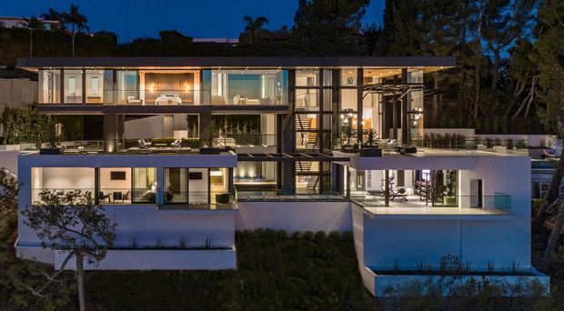 Twilight exterior view of a large contemporary multi-level residence on a sloped, landscaped site. The home features extensive floor-to-ceiling glazing, broad flat roof planes, projecting terraces, white rendered volumes, dark structural framing, and warmly illuminated interior spaces visible through the glass.