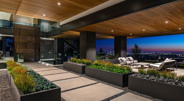 A sophisticated rooftop terrace or elevated outdoor entertaining area photographed at dusk. Large concrete pavers with dark gravel joints form a walkway between raised black planter boxes filled with ornamental grasses and succulents. A deep timber-lined ceiling with recessed lighting is supported by substantial dark structural beams and columns. Contemporary outdoor lounge furniture overlooks a broad cityscape, with extensive glazing and dark framed architectural elements connecting the terrace to the interior.