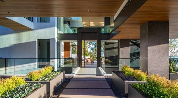 Contemporary multi-level building entry viewed from a landscaped approach, with broad glazed doors, glass balustrades, dark masonry or rendered columns, and a warm timber-lined soffit. Rectangular raised planter beds and a paved walkway frame the entrance, while an open interior atrium is visible beyond the doors.