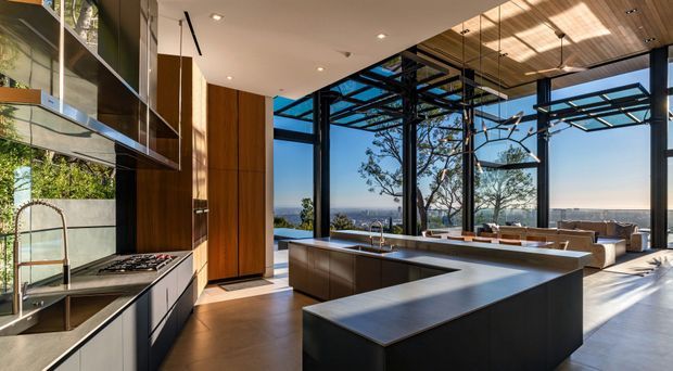 A spacious contemporary open-plan kitchen with a large L-shaped island and integrated sink, dark cabinetry, timber wall joinery, and expansive black-framed floor-to-ceiling glazing. The kitchen opens directly to a bright living area and covered outdoor terrace, with a view toward a blue coastal or ocean-like horizon. A timber slatted ceiling and exposed dark structural framing add architectural interest.