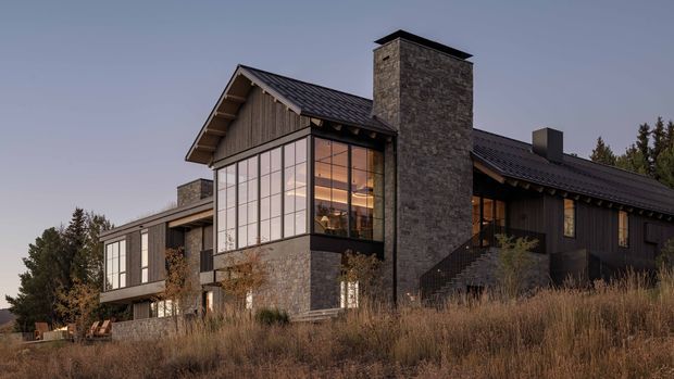 Exterior view of a substantial contemporary mountain-style residence set in a grassy, wooded landscape. The house features dark pitched roof forms, extensive floor-to-ceiling glazing, dark vertical timber cladding, natural stone walls, and a prominent central stone chimney. Warm interior lighting is visible through the glazed living spaces, creating a refined evening atmosphere.