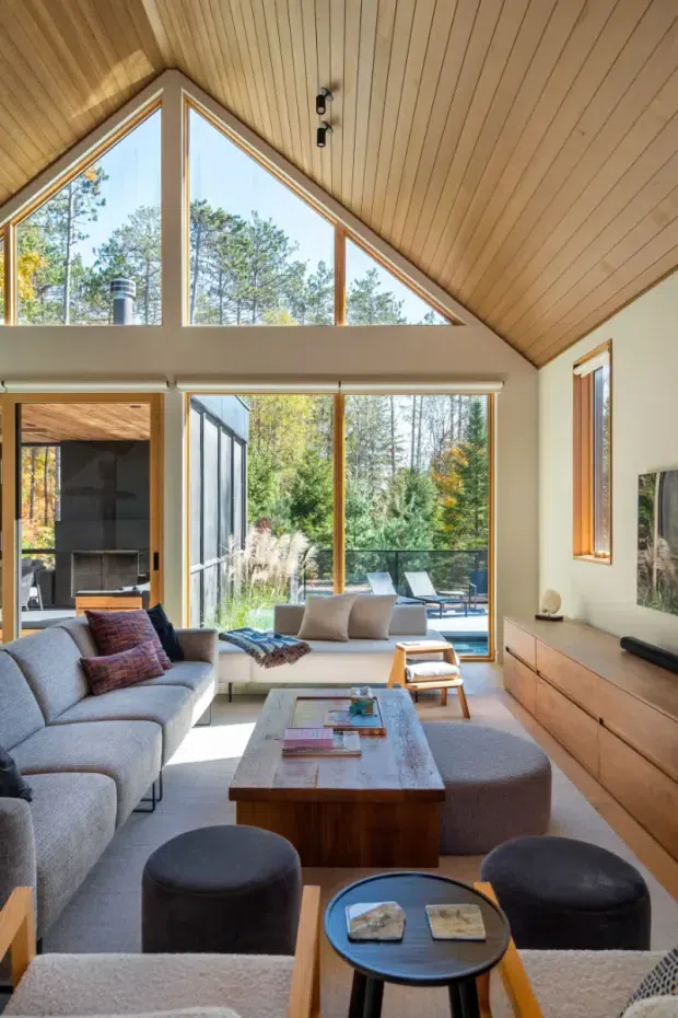 A bright, contemporary living room with a dramatic vaulted timber-lined ceiling and large triangular and floor-to-ceiling windows overlooking a wooded landscape. The room includes a large pale sectional sofa, upholstered ottomans, a low timber coffee table, built-in timber media cabinetry, and a compact sitting area by the glazing. Warm natural wood, abundant daylight, and strong indoor-outdoor connection define the space.