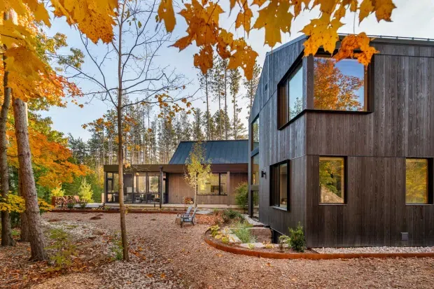 Exterior view of a contemporary multi-volume home set within a wooded landscape during autumn. The building features dark vertical timber cladding, large framed windows, a steeply pitched metal roof on the central volume, and a crisp box-like two-storey wing connected by glazed sections. A gravel courtyard and curved planted garden bed occupy the foreground.