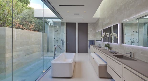 Contemporary, spacious bathroom with a freestanding white bathtub beside full-height glazing, a long double vanity with pale cabinetry and integrated sinks, and a large illuminated mirror. Light stone or porcelain wall cladding, pale flooring, recessed ceiling lighting, and views to a planted outdoor area create a bright, refined atmosphere.