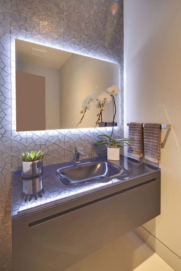 Compact contemporary bathroom vanity with a floating taupe-grey cabinet, integrated dark glossy countertop and basin, geometric metallic-grey wall tiles, and a large backlit mirror. A chrome tap, folded towels on a wall-mounted rail, and potted orchids and succulent plants add detail. The space is softly illuminated with a refined, spa-like atmosphere.