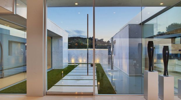 A refined contemporary interior looking through full-height glazed doors and walls onto a narrow landscaped courtyard with large pale stepping slabs, clipped grass strips, and a waterfront or pool-like body of water beyond. The interior has polished light flooring, white walls, recessed ceiling lights, extensive glass, and sculptural dark decorative objects displayed on plinths. The view is symmetrical and minimalist, with strong indoor–outdoor visual connection and evening light.