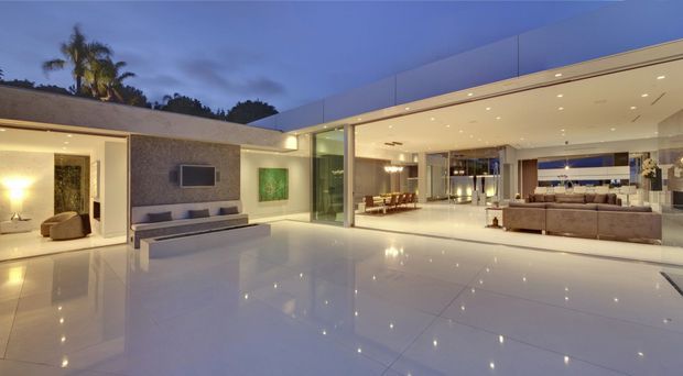 Wide dusk view of a luxurious contemporary open-plan residence, showing a glossy white tiled courtyard or living area opening directly into a bright living room and dining space. The building has clean rectilinear forms, extensive floor-to-ceiling glazing, a flat roof, recessed downlighting, and a restrained neutral palette. A textured feature wall with wall-mounted television, low built-in joinery, sofas, dining furniture, and tropical planting are visible.