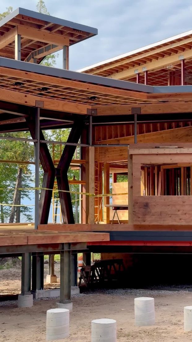 Photograph of a multi-level building under construction, showing exposed timber framing, engineered wood beams and decking, dark steel structural columns and bracing, open floor slabs, and concrete pier footings. The structure has broad projecting roof planes and a partially elevated form set among trees.