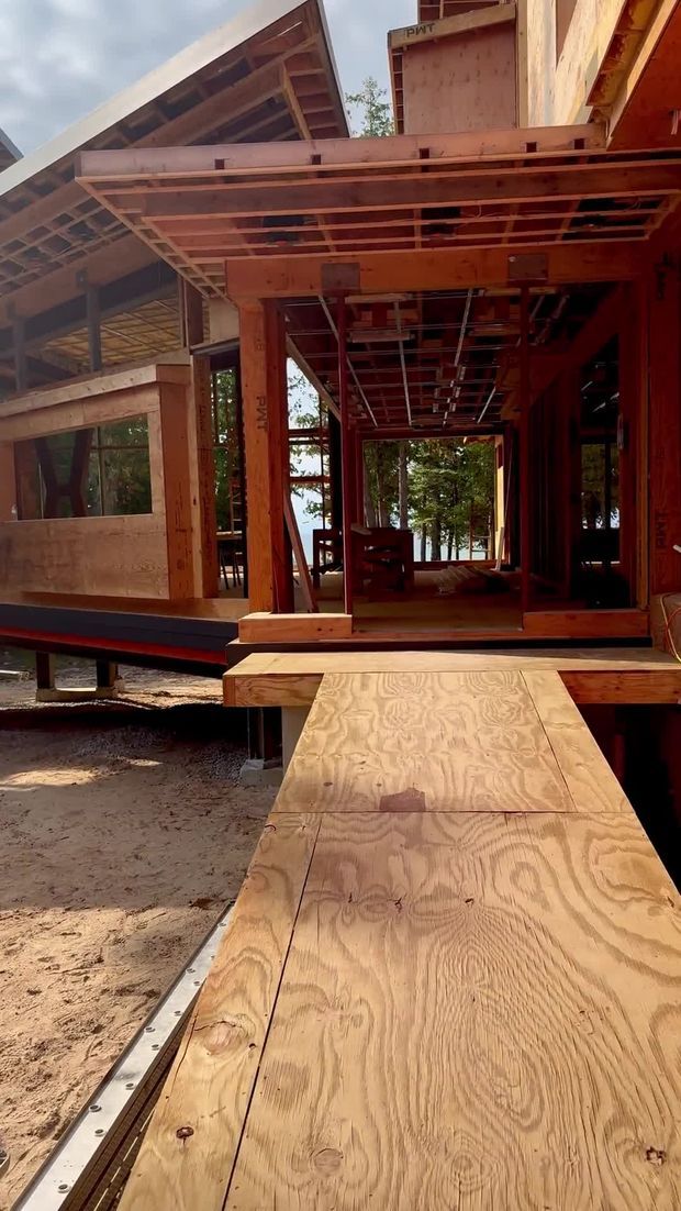 Photograph of a timber-framed building under construction, viewed from an unfinished plywood access deck or ramp. Exposed structural wood framing, plywood sheathing, roof framing, open wall cavities, and temporary supports are visible. The structure appears elevated above the ground, with trees and a bright body of water visible beyond the open interior.