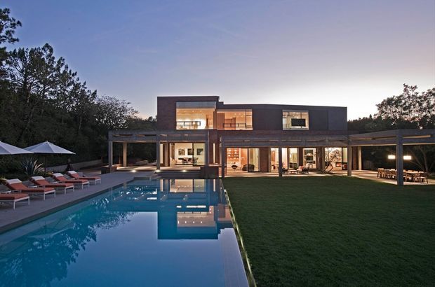 Twilight exterior view of a large contemporary two-storey residence with a rectilinear dark-clad facade, extensive floor-to-ceiling glazing, covered outdoor living areas, and a reflecting swimming pool in the foreground. The home is set within landscaped grounds and mature trees, with illuminated interiors visible through the glazing.