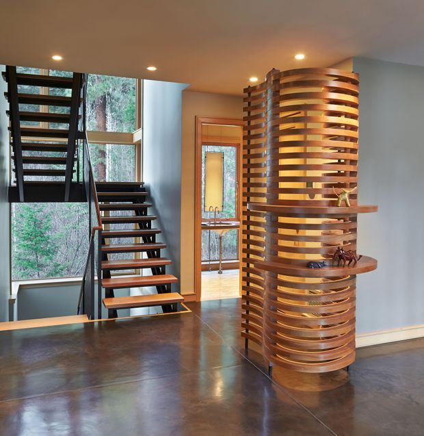 Interior view of a contemporary entry hall with an open-riser staircase, expansive forest-facing glazing, polished concrete flooring, and a striking curved cylindrical timber slat screen with integrated display shelves. Warm recessed lighting contrasts with cool grey walls and dark stair structure.