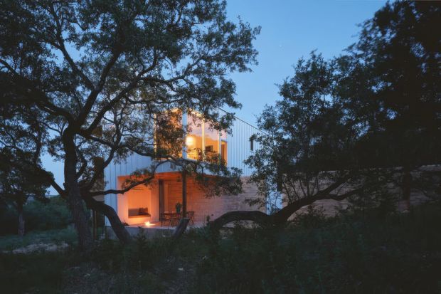 Twilight exterior view of a contemporary multi-level residence partially concealed by mature trees and dense landscaping. The home features crisp rectilinear volumes, light-colored vertical metal cladding, warm timber-toned wall surfaces, and strongly illuminated glazed openings and outdoor areas, creating a striking contrast against the dark natural setting.