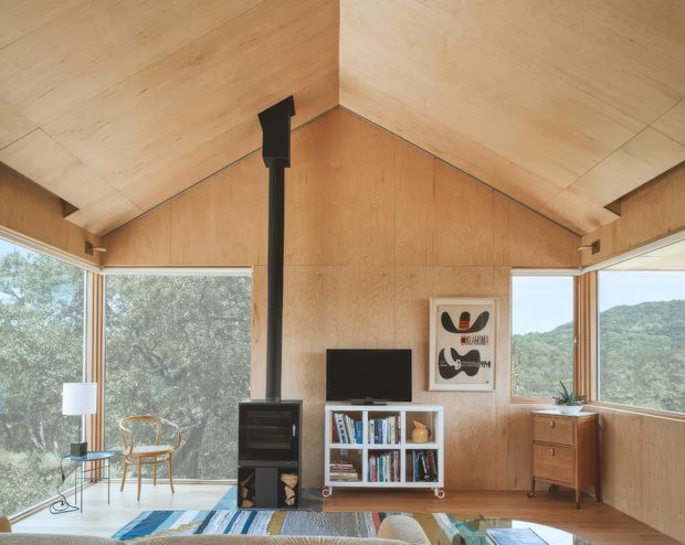 A compact, wood-lined living room with a dramatic pitched ceiling and exposed plywood wall and ceiling surfaces. Large corner windows bring in abundant natural light and frame leafy hills beyond. A black freestanding wood-burning stove, television, low bookshelf, mid-century-style timber cabinet, framed artwork, chair, and patterned rug furnish the room. The space has a warm cabin-like, minimalist character with strong indoor-outdoor connection.