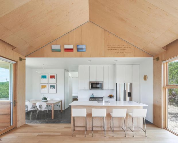 Bright open-plan kitchen and dining area within a timber-lined room featuring a high pitched ceiling. The kitchen has white full-height cabinetry, a central island with seating for four, stainless-steel appliances, and a pale benchtop. A dining table is visible beyond the island, with large timber-framed windows and glazed doors bringing in abundant natural light. Small artwork and wall text decorate the timber gable wall.