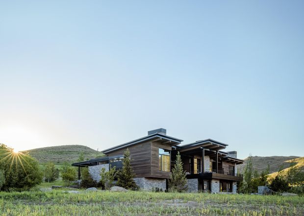 A contemporary multi-level house set in an open, hilly landscape. The building has extensive horizontal timber cladding, dark metal roof planes and window frames, stone-clad lower walls, deep roof overhangs, and large areas of glazing. Low native-looking landscaping surrounds the house, with the low sun visible behind the hills.