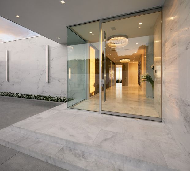 A refined modern building entrance with broad pale stone steps and matching marble-look wall cladding. Large frameless glass pivot doors open into a bright, elongated foyer with polished light flooring, warm recessed lighting, decorative circular chandeliers, and a plant. A minimalist landscaped strip and covered entry canopy frame the approach.