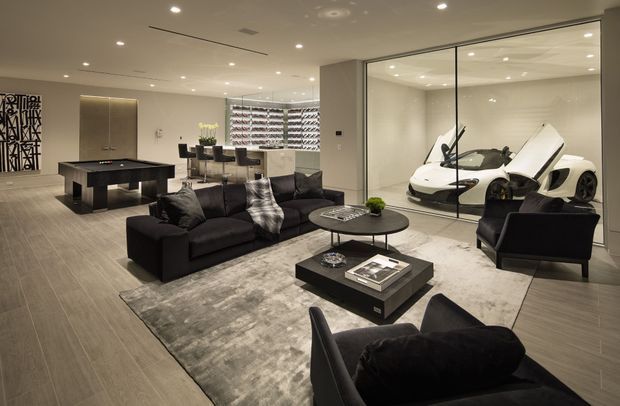 A sophisticated open-plan luxury living room with a dark sectional sofa, black armchairs, layered coffee tables, and a large muted area rug. The space includes a dining area and compact kitchen-style joinery in the background, while a fully glazed internal display garage showcases two white sports cars. Recessed ceiling lighting, pale timber-look flooring, neutral walls, and dark furnishings create a refined contemporary showroom-like atmosphere.