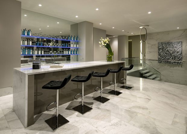 Luxurious contemporary bar and entertaining area with a long white marble island, four black bar stools, mirrored shelving displaying bottles, recessed ceiling lights, and a polished marble floor and wall. A small stepped transition and decorative artwork are visible in the background.