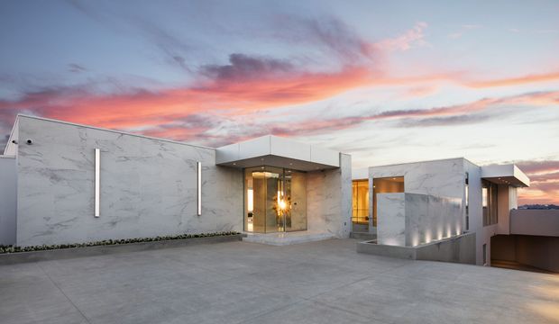 Contemporary low-profile residence photographed from a broad concrete driveway or forecourt at sunset. The house features a strongly horizontal composition, pale marble-look exterior wall surfaces, projecting flat roof forms, recessed warm lighting, large glazed entry doors, and a lower-level garage opening at the right. The dramatic pink and blue sky is visible above the building.