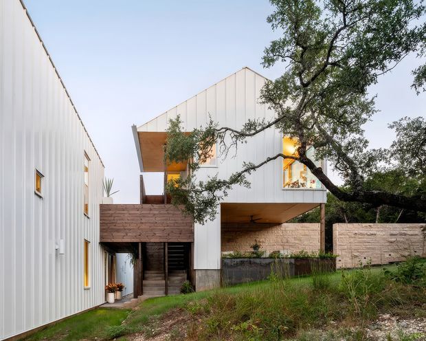 Exterior view of a contemporary multi-level residence integrated into a wooded, sloped site. The building features white vertically ribbed cladding, steep gabled roof forms, warm timber-lined soffits and recessed balconies, with exposed masonry retaining walls and large illuminated windows. Mature trees and natural grasses frame the structure.