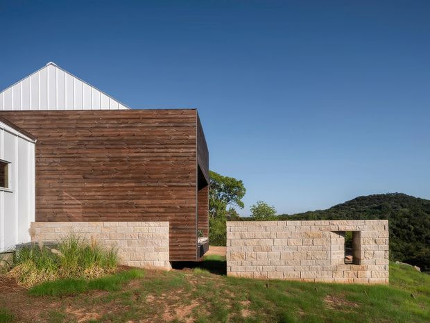 Exterior architectural view of a contemporary building composed of white vertical metal cladding and a projecting timber-clad volume, with a low pale masonry wall and rectangular opening in the foreground. The structure sits on a grassy site with wooded hills in the background beneath a clear blue sky.