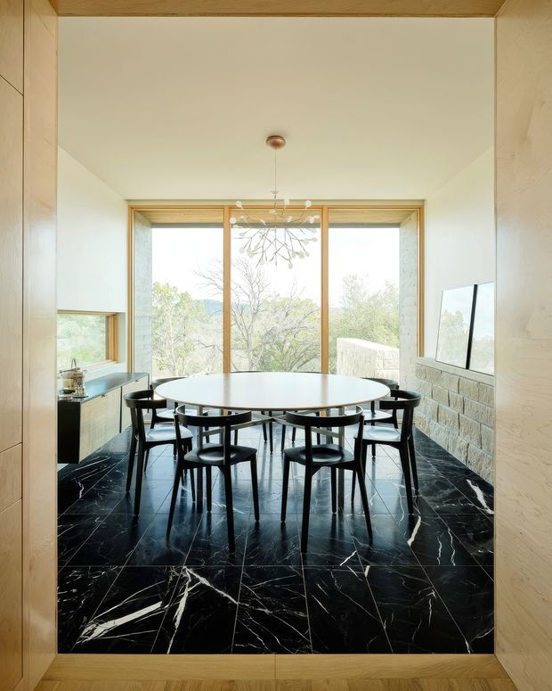 A refined dining room viewed through a timber-framed opening, featuring a large round white dining table surrounded by black sculptural chairs. The room has dramatic black marble-look flooring with prominent white veining, pale textured walls, warm timber framing and cabinetry, expansive floor-to-ceiling glazing, and a delicate contemporary pendant light. A stone-clad feature wall and framed artwork are visible on the right, with leafy outdoor views beyond the windows.