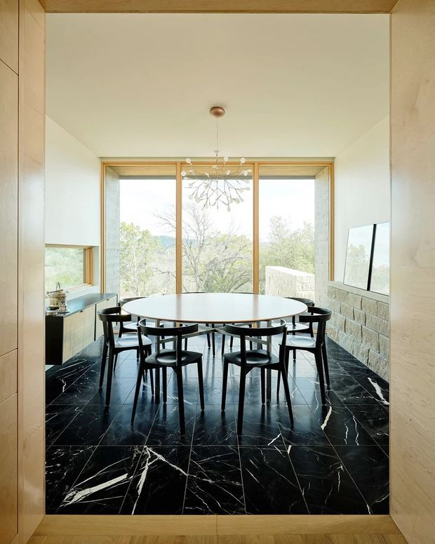 A refined dining room viewed through a timber-framed opening, featuring a large oval dining table surrounded by black sculptural chairs. The room has dramatic black marble-look floor tiles with white veining, pale plaster walls, warm timber window and doorway frames, a pale stone masonry feature wall, expansive glazing, and a delicate branching pendant light. A compact kitchen or kitchenette is visible to the left.