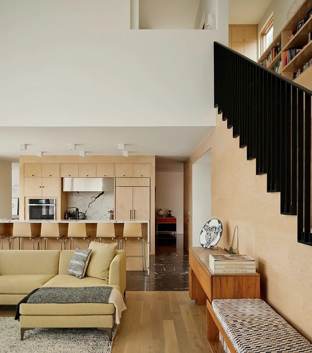 A warm, refined open-plan living space with a compact kitchen and island in the background, a pale upholstered sectional sofa in the foreground, and a timber console with a patterned bench along the wall. Pale timber kitchen cabinetry and timber flooring contrast with a dramatic black vertical-slat stair balustrade. The space has a double-height or partially open stair volume, built-in shelving, neutral plaster walls, and a dark stone or tile transition floor near the kitchen.