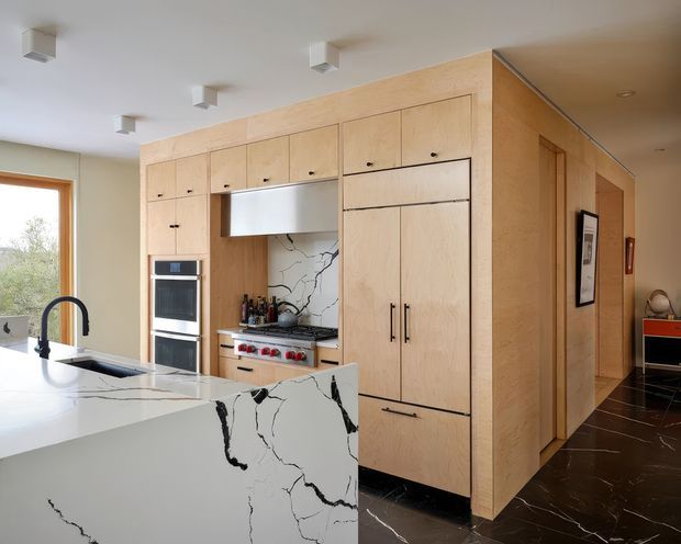 A bright contemporary kitchen with a large white island and integrated sink, black gooseneck tap, pale timber cabinetry, built-in oven, concealed rangehood and marble-look splashback and benchtops with dramatic dark veining. The kitchen opens toward a hallway, with dark veined flooring and minimal white ceiling lights.