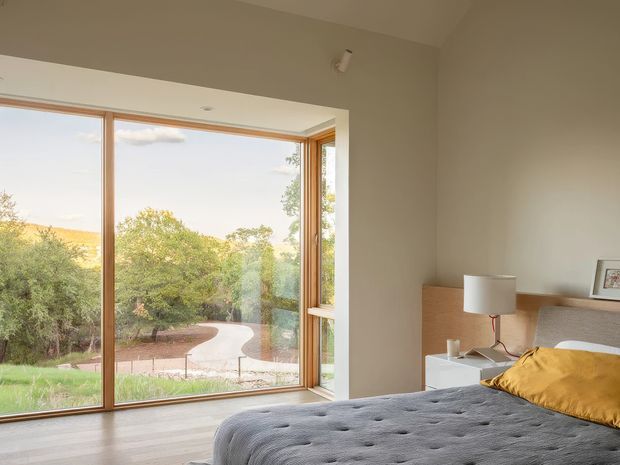 A bright, contemporary bedroom with a large corner window providing expansive views of a landscaped garden and curved pathway. The room has pale neutral walls, light timber flooring, a low timber bedside unit, a white table lamp, framed artwork, and a grey upholstered bed with a mustard-yellow accent cushion. The glazing and warm timber frames create a strong connection to the outdoors.