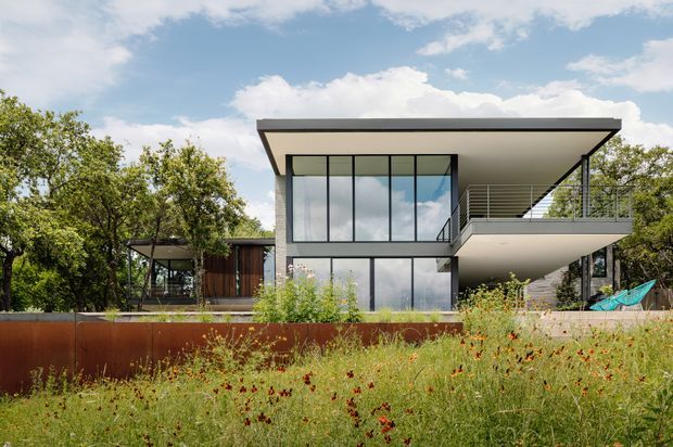 A modern two-storey residential house set within a landscaped, meadow-like garden. The building has a strongly horizontal flat roof, extensive floor-to-ceiling glazing, dark slender window frames, a projecting upper-level balcony, and a mix of pale rendered surfaces and warm timber elements. A rusted steel retaining wall runs across the foreground, with mature trees surrounding the home.