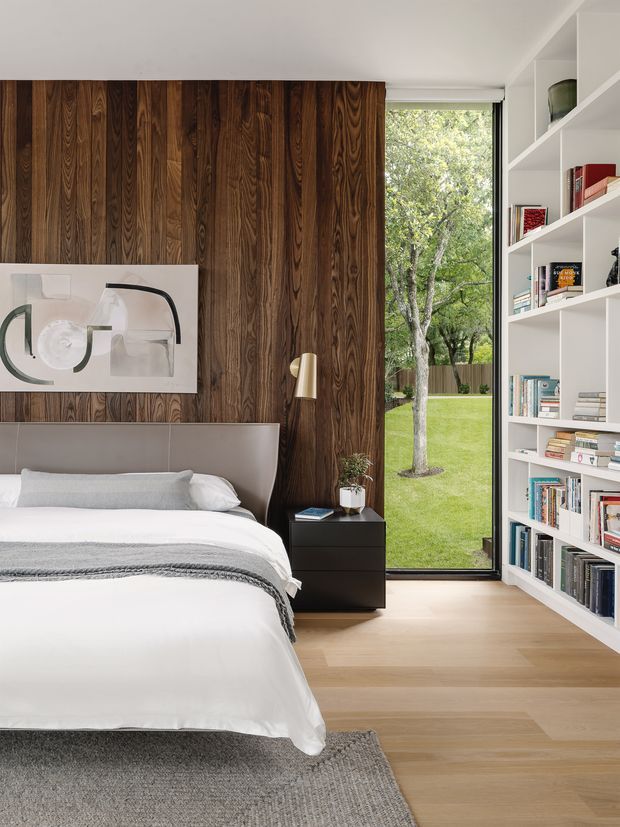A refined contemporary bedroom with a partially visible bed dressed in white linens and a grey throw, set against a dramatic dark timber feature wall. A black bedside table and slim wall sconce sit beside the bed, while full-height glazing provides a view to a green garden. Floor-to-ceiling white built-in shelving filled with books occupies the right side, and light timber flooring with a grey rug completes the room.