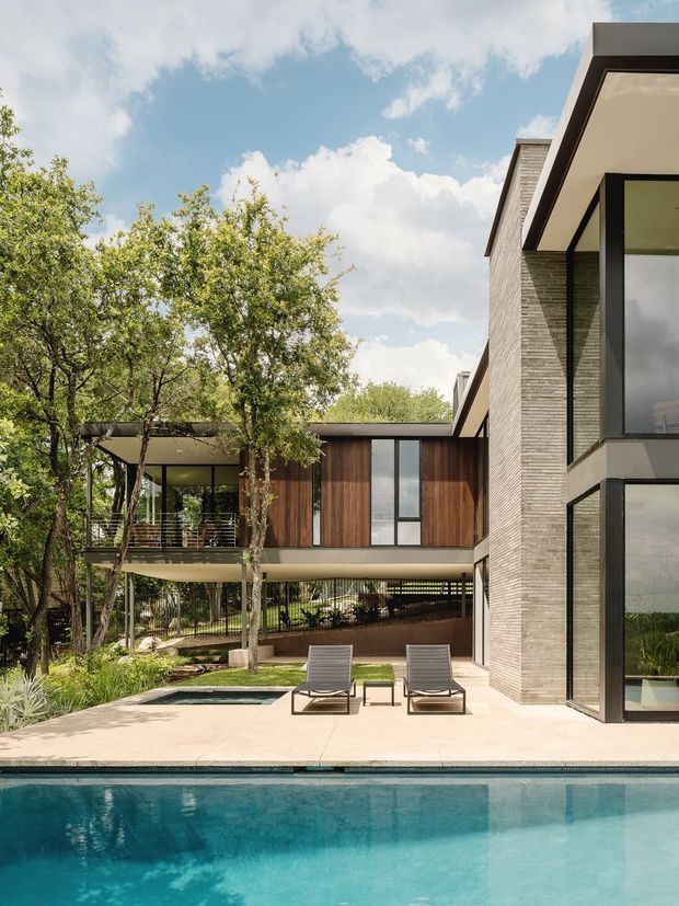 A contemporary multi-level residence viewed from the pool terrace, featuring a flat-roofed composition, expansive black-framed glazing, pale textured masonry walls, warm timber cladding, and covered outdoor living areas integrated into a landscaped courtyard. Two loungers sit beside the swimming pool in the foreground.