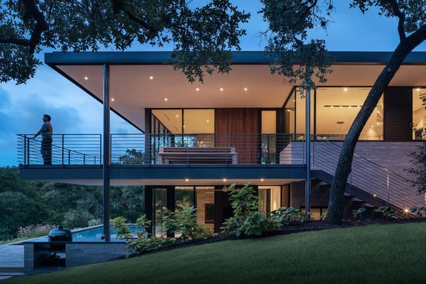 A contemporary two-storey house viewed from the landscaped rear or side yard at dusk. The home features a broad flat roof, extensive floor-to-ceiling glazing, slim dark metal framing, open upper-level terraces with horizontal metal railings, warm recessed lighting, timber-toned interior surfaces, and a projecting balcony. Mature trees and sloping lawn frame the structure, with a built-in outdoor grill or kitchen element visible at lower left.