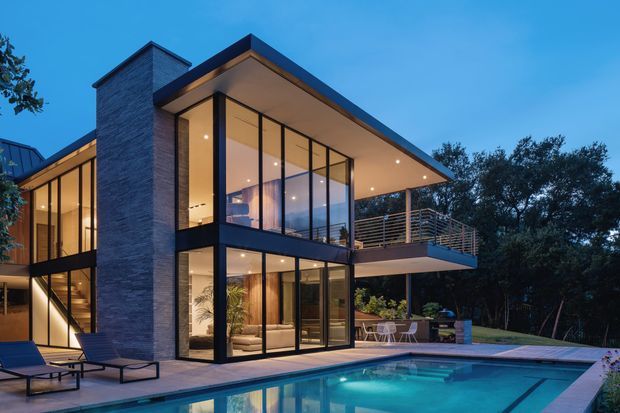 Twilight exterior view of a contemporary multi-level house with a flat projecting roof, tall floor-to-ceiling glazing, dark framed windows, a tall stacked-stone feature wall, and a cantilevered upper balcony. A swimming pool and paved terrace occupy the foreground, with warm interior lighting visible through the glazing and landscaped trees surrounding the property.