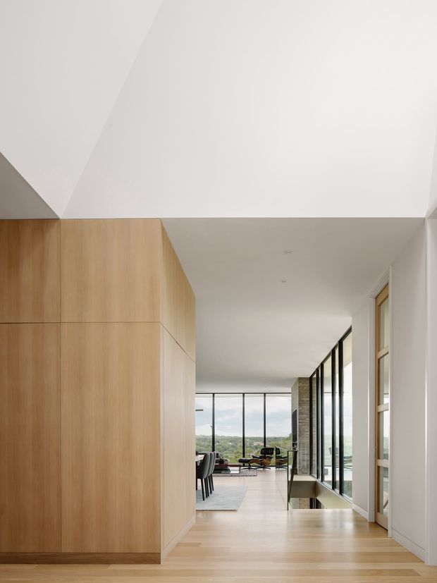 A bright, minimalist open-plan interior viewed along a timber-lined corridor toward a dining and living area. Full-height black-framed glazing opens to a green landscape, while pale timber flooring, integrated timber cabinetry, white walls, and a sculptural angular ceiling create a clean contemporary feel.
