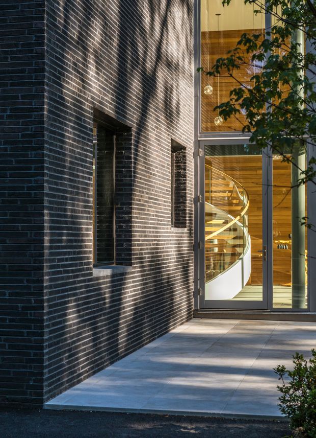 Exterior architectural photograph of a contemporary building entrance. A dark charcoal brick facade with narrow vertical windows frames a paved entry terrace, while a tall glazed doorway reveals a warm timber-lined interior and a sculptural white spiral staircase. Grey metal window and door frames, strong sunlight, and partial landscaping complete the scene.